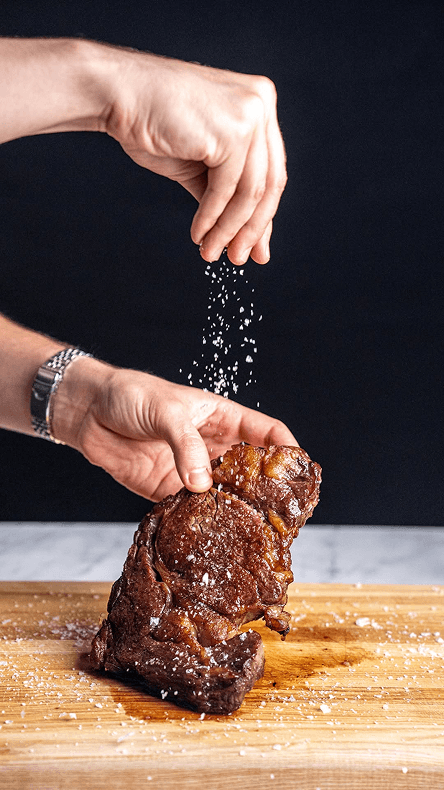 Hands sprinkling salt onto a steak resting on a wooden board